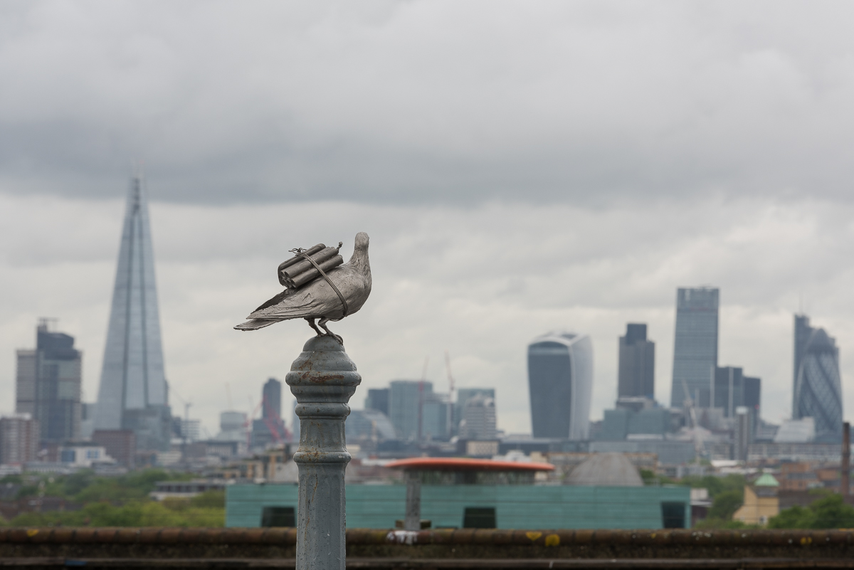 ID: a life-sized bronze cast of a pigeon with a stick of dynamite and blackberry phone on its back is placed on a London bollard looking out toward the London skyline with ominous clouds above.