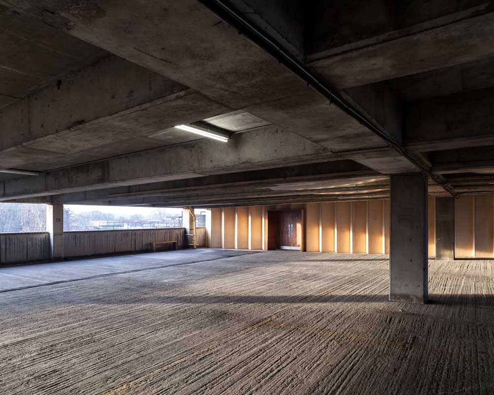 ID: a wide angle view of the architecture of a single level of a brutalist car park. At the back of the level there is a concertina wall that stretches its entire width and is made from light wood with several in-set double doors.