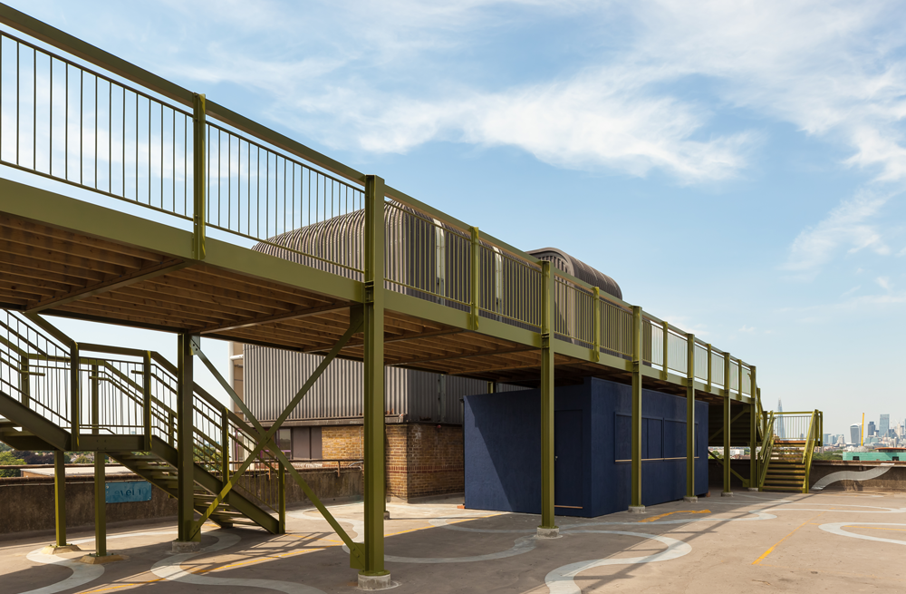 ID: a large steel platform is installed on top of a concrete rooftop. It is painted soft green and its architecture is straight and minimalistic and mirrors that of the original car park. Beautiful blue skies are seen in the background.