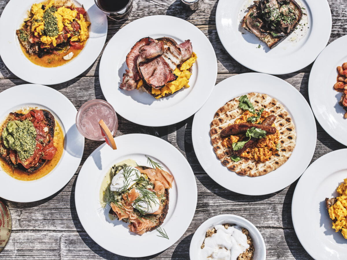 ID: a bird's eye view of 6 plates of brunch food on white plates on a bleached wooden surface. The food includes bacon and scrambled egg, salmon and poached eggs and flatbread and humous.