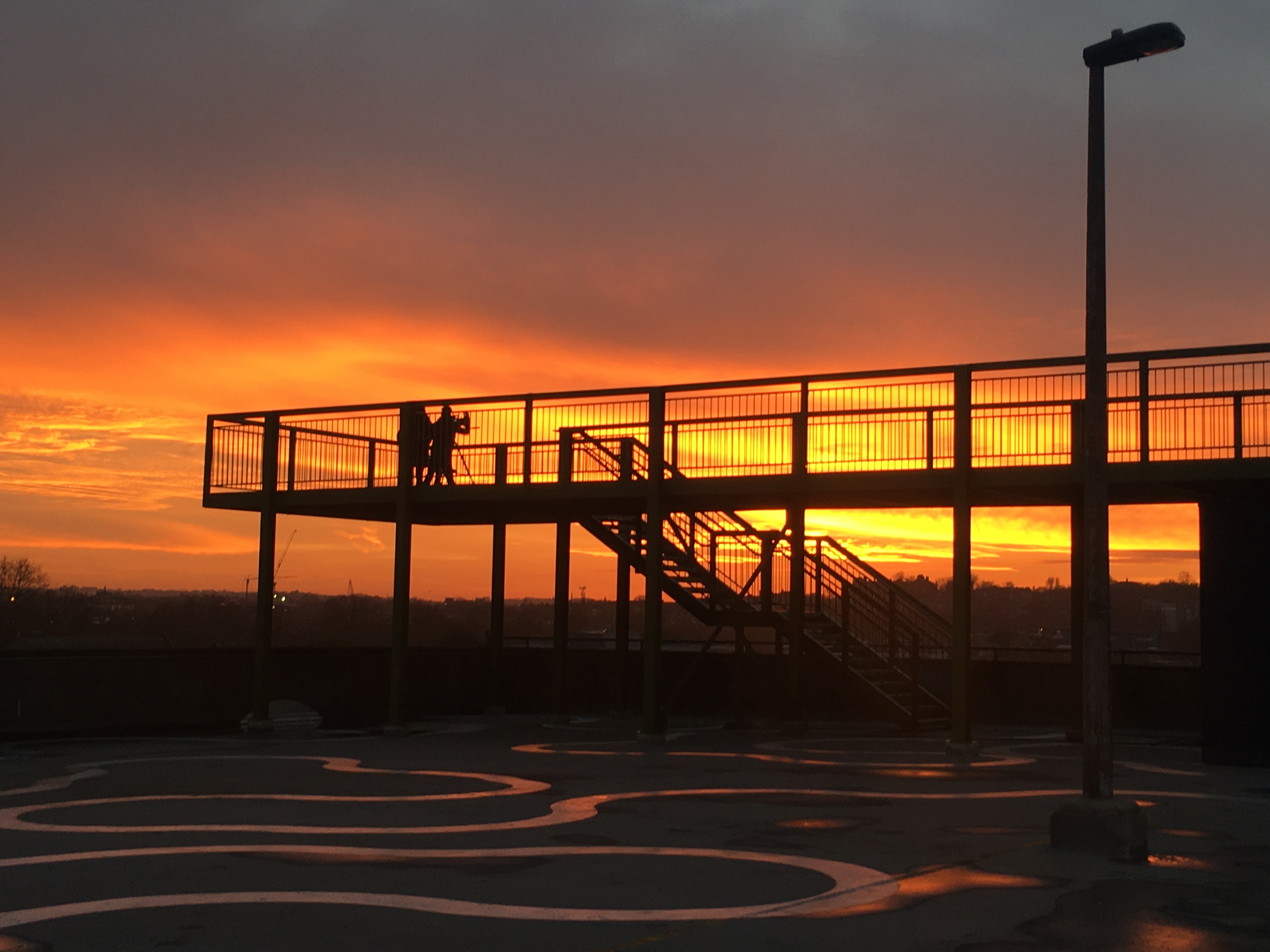 The silhouette of a large steel platform that stands on an expansive concrete rooftop is set against a vivid and warm orange sunset.