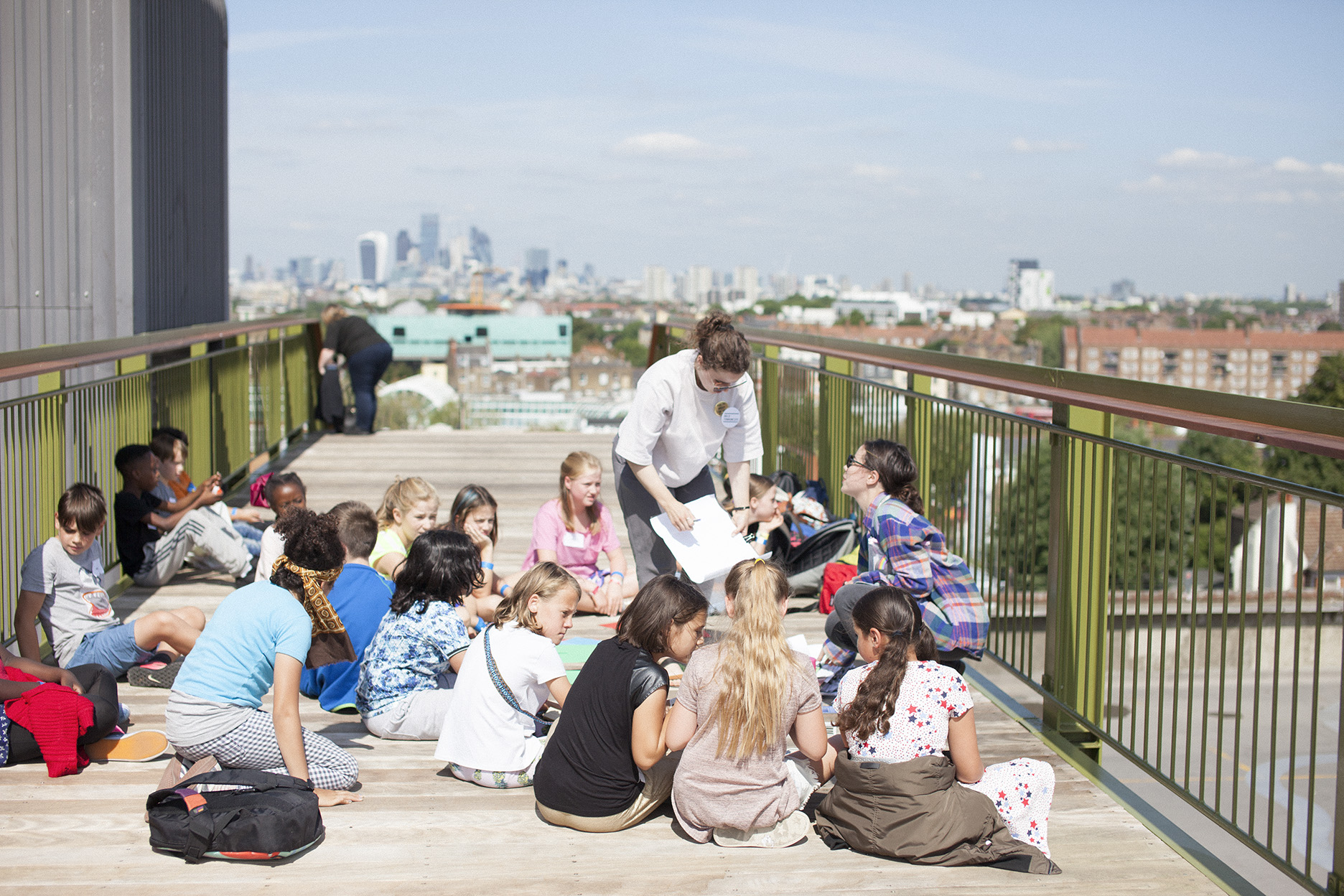 ID: a group of 15 young school children are sat on a raised platform on a concrete rooftop with the London skyline and blue skies in the background. They are sketching in notebooks and taking instructions from an adult workshop leader.
