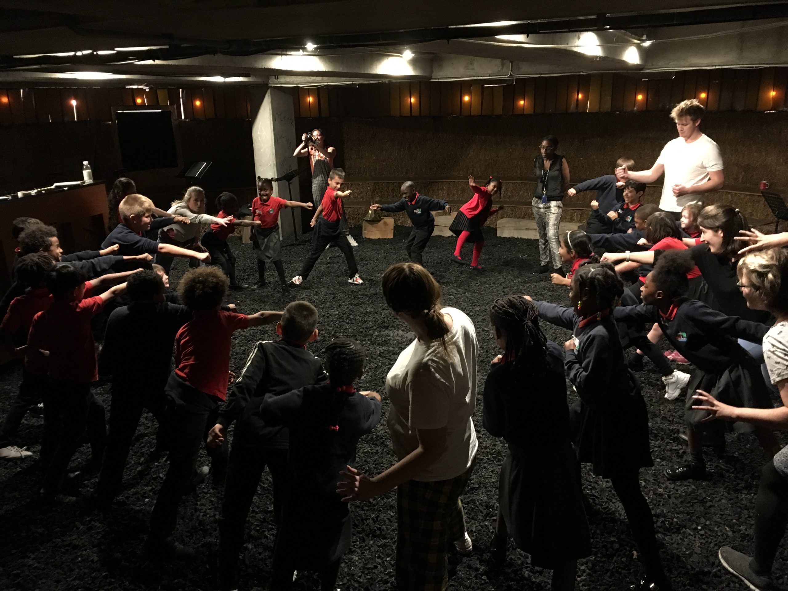 ID: a full classroom of young children are stood in a circle inside a concrete room with rubber flooring, making an active pose. They are led by an adult workshop leader.