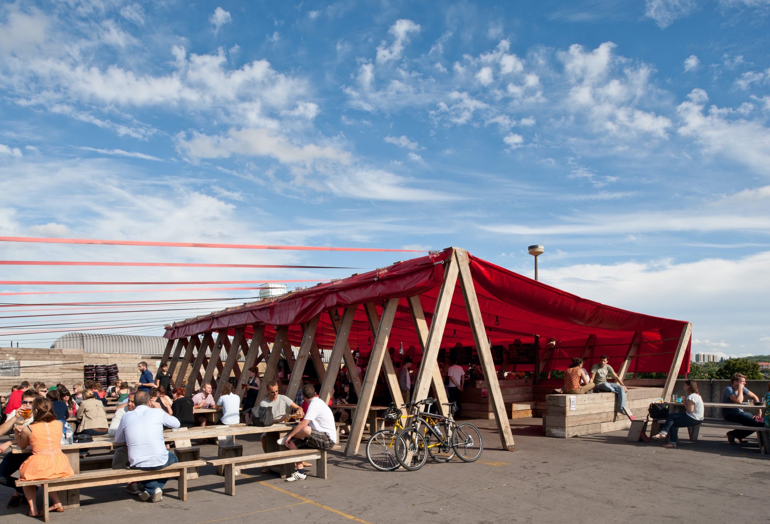 ID: a large wooden A-frame is used to support a large red canopy over a rooftop cafe and restaurant. People are seen sitting on tables in the foreground and enjoying refreshments.