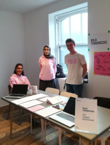 ID: three young adults wearing pink Bold Tendencies apparel are stood behind a desk at a careers fair. On the table is an open laptop and lots of different sign up sheets and pamphlets. 