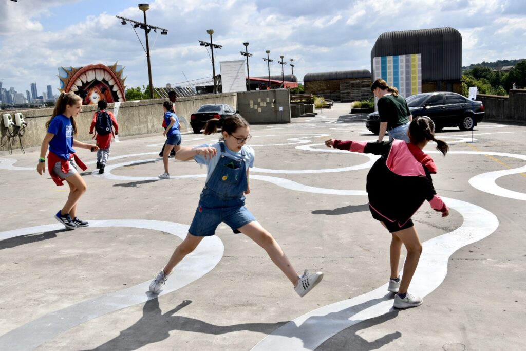 ID: a number of young school children are jumping between silver curved lines painted directly onto a concrete rooftop. In the background there are a number of colour artworks installed on the rooftop.