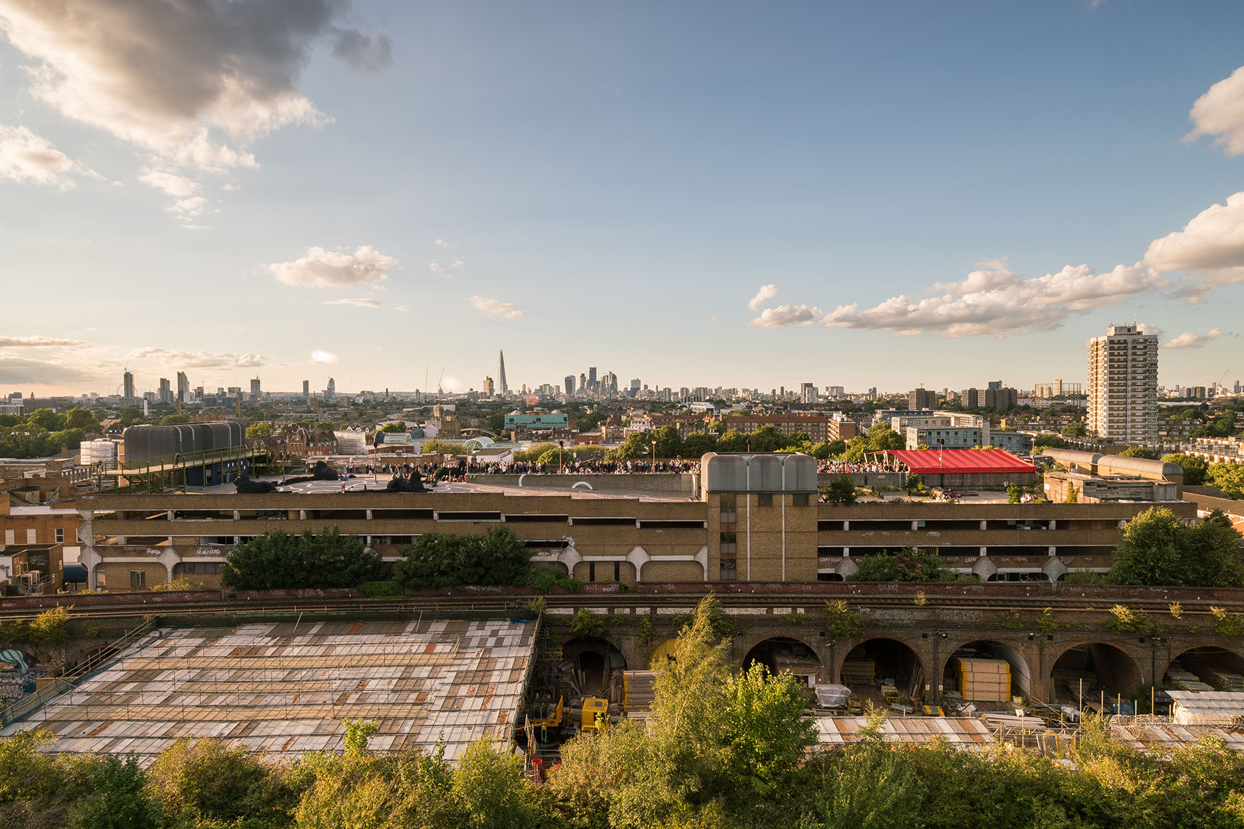 ID: A wide angle view taken from a great height looks out toward the side profile of a brutalist multi-storey car park. On the rooftop there are people walking around and large artworks. Beyond the rooftop you can see the full London skyline set against clear blue skies.