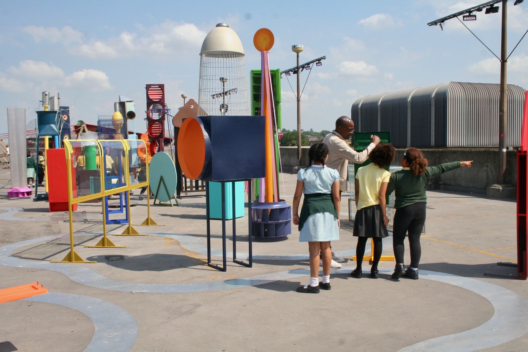 ID: three young school children are escorted through a colourful art installation made from repurposed and painted waste products. The installation is on top of a concrete rooftop with clear blue skies in the background.