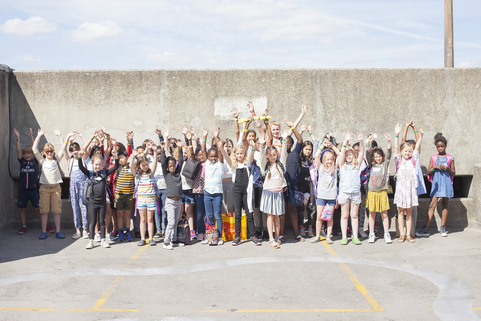 ID: a full classroom of 30 young school children and their teachers stand in front of a concrete wall on a large rooftop with their hands in the air and smiling.