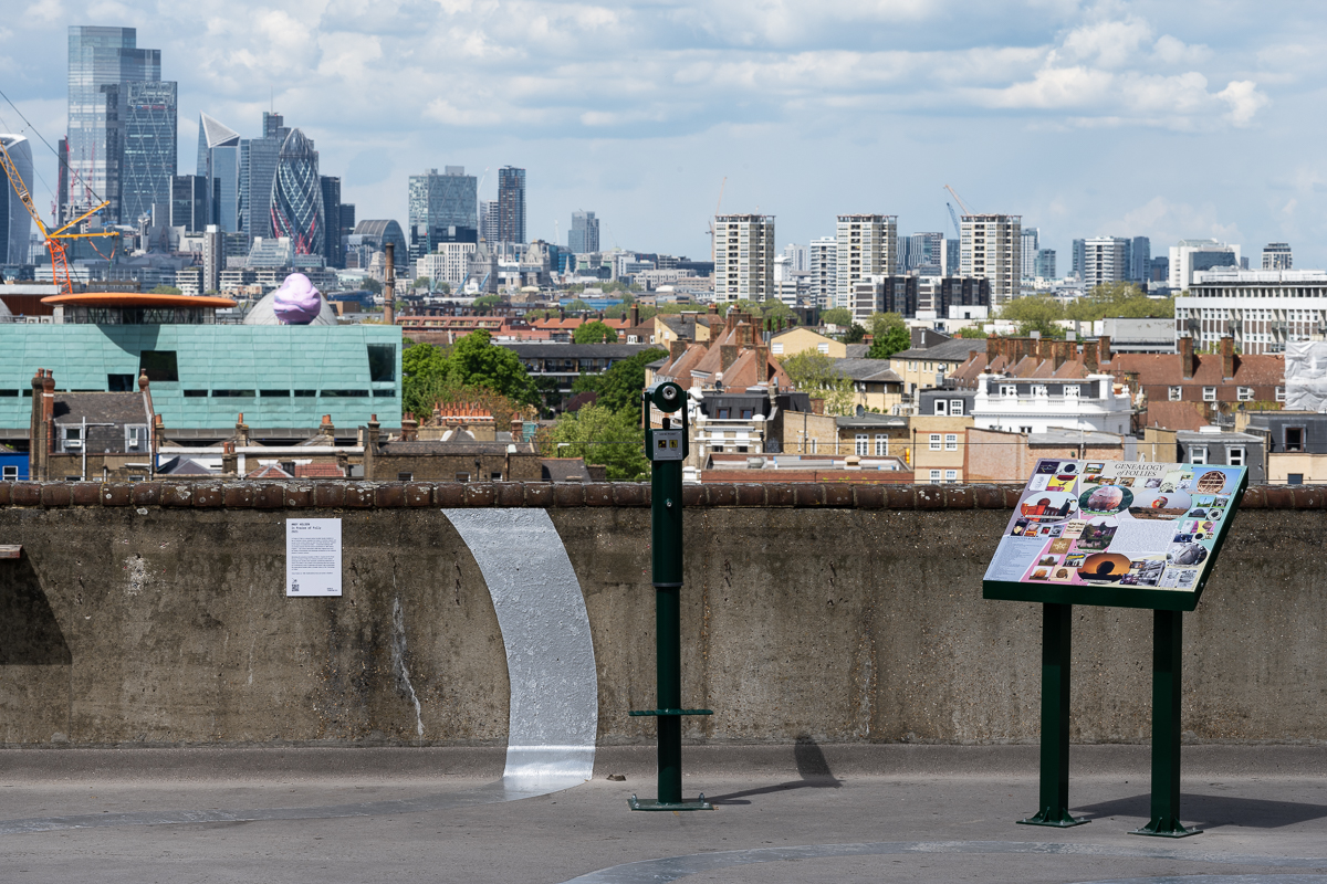 ID: a view of the London skyline from South London. In the centre is Peckham Library and on top of this building is installed a 5m tall pink and purple boulder. In the foreground, a green tourist telescope and information lectern are displayed on a concrete rooftop, approximately 400m away.