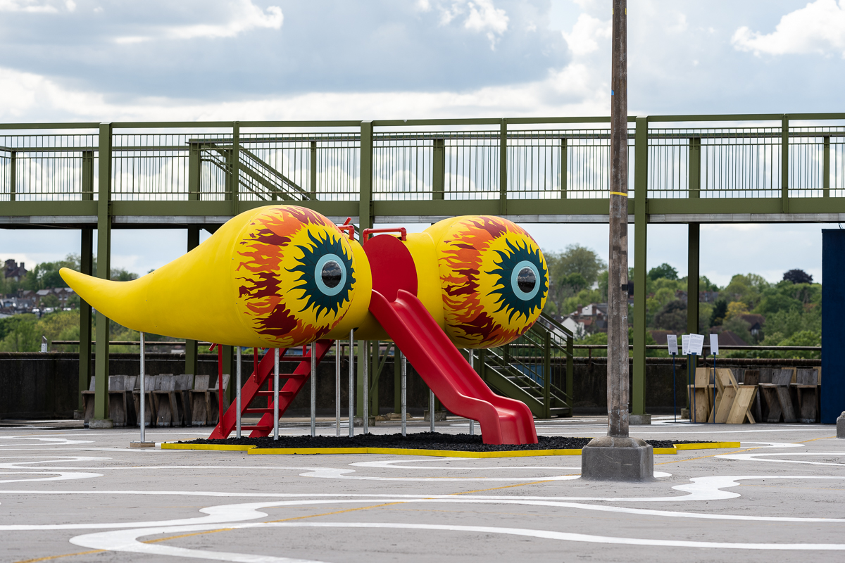ID: a bright yellow spaceship with a red playground slide, central platform and steps is installed on a concrete rooftop. The spaceship is raised 1.5 meters from the ground to a total height of 3.5 metres and is approximately 4.5 metres long and wide. At the front of the ship are two dome-shaped eyes that tail into long tadpole shapes toward the rear. Each dome is hand-painted with flames in a mixture of green, turquoise and orange, and a transparent iris at their centre emits a beam of light from within the vessel. The spaceship is surrounded by a pit of jet-black rubber mulch contained within a diamond perimeter painted in matching yellow.