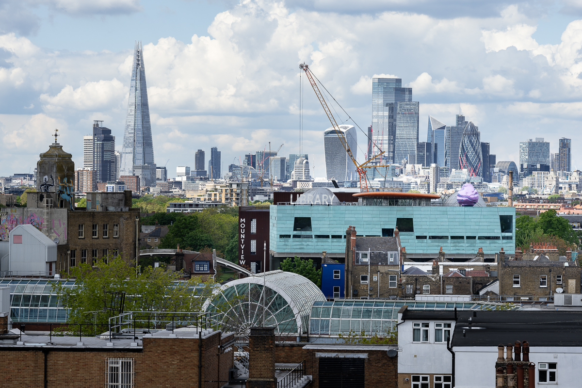 ID: a view of the London skyline from Peckham stretching from the shard on the left hand side to the gherkin on the right hand side. In the centre is Peckham Library and on top of this building is installed a 5m tall pink and purple boulder.