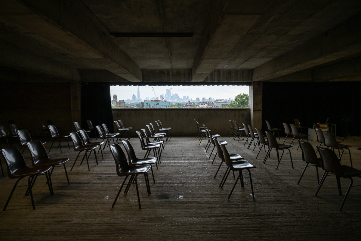 ID: rows of brown chairs are placed inside a covered space in a concrete car park, facing tower the right hand side of the image. A window section of the back wall looks out toward the London skyline.