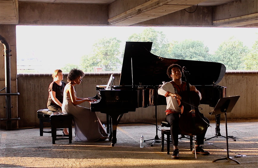 ID: two artists, one on a grand piano, the other on cello, perform inside a covered concrete area. A window in the background shows green foliage and strong sun.