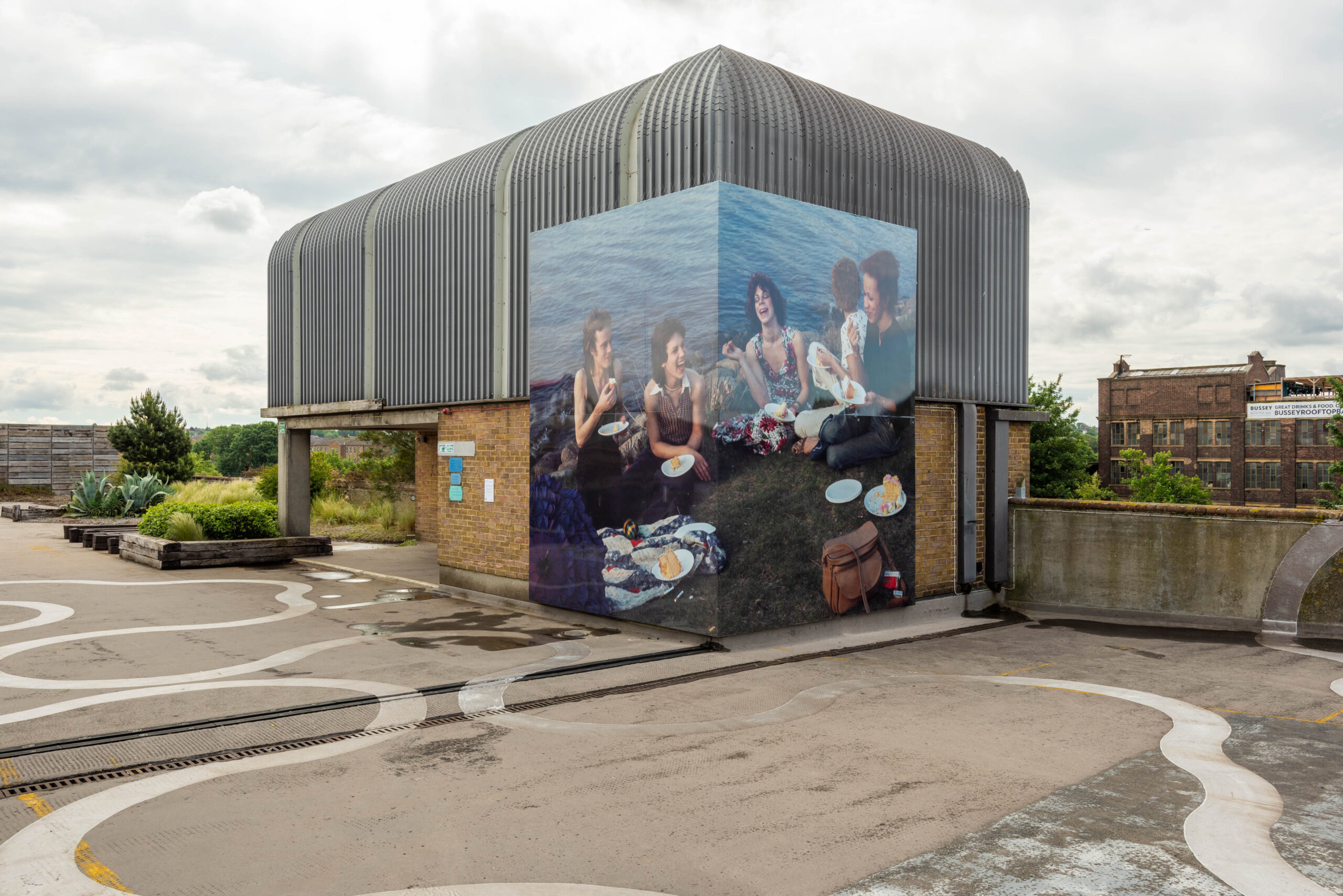 ID: a large billboard is attached to a rooftop lift shaft, measuring 4 metres tall by 6 metres wide. There is a right angle bend half way through the width of the image as it wraps around the lift shaft. The image shows four people sitting on the grassy bank of a clear river, dressed in jeans, t-shirts and flowery summer dresses. There is a parasol, bottle, cigarette packet and a camera case on the floor around them. They are all joyfully laughing as they eat slices of sponge cake from white paper plates.