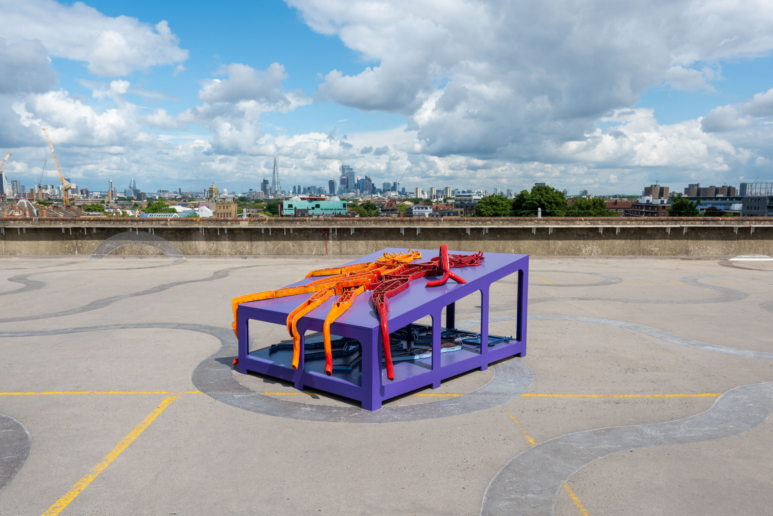 ID: a bright purple structure in the shape of a large tomb with two levels and a row of hollow arches on either side is placed on a concrete rooftop with the London skyline and blue skies in the background. On the top level of the structure two figures are made out of red and orange ceramic, overlapping anatomies and colours. On the bottom level, a pair of skeletons are made from sections of blue ceramic, mirroring the relationship of the top two figures.