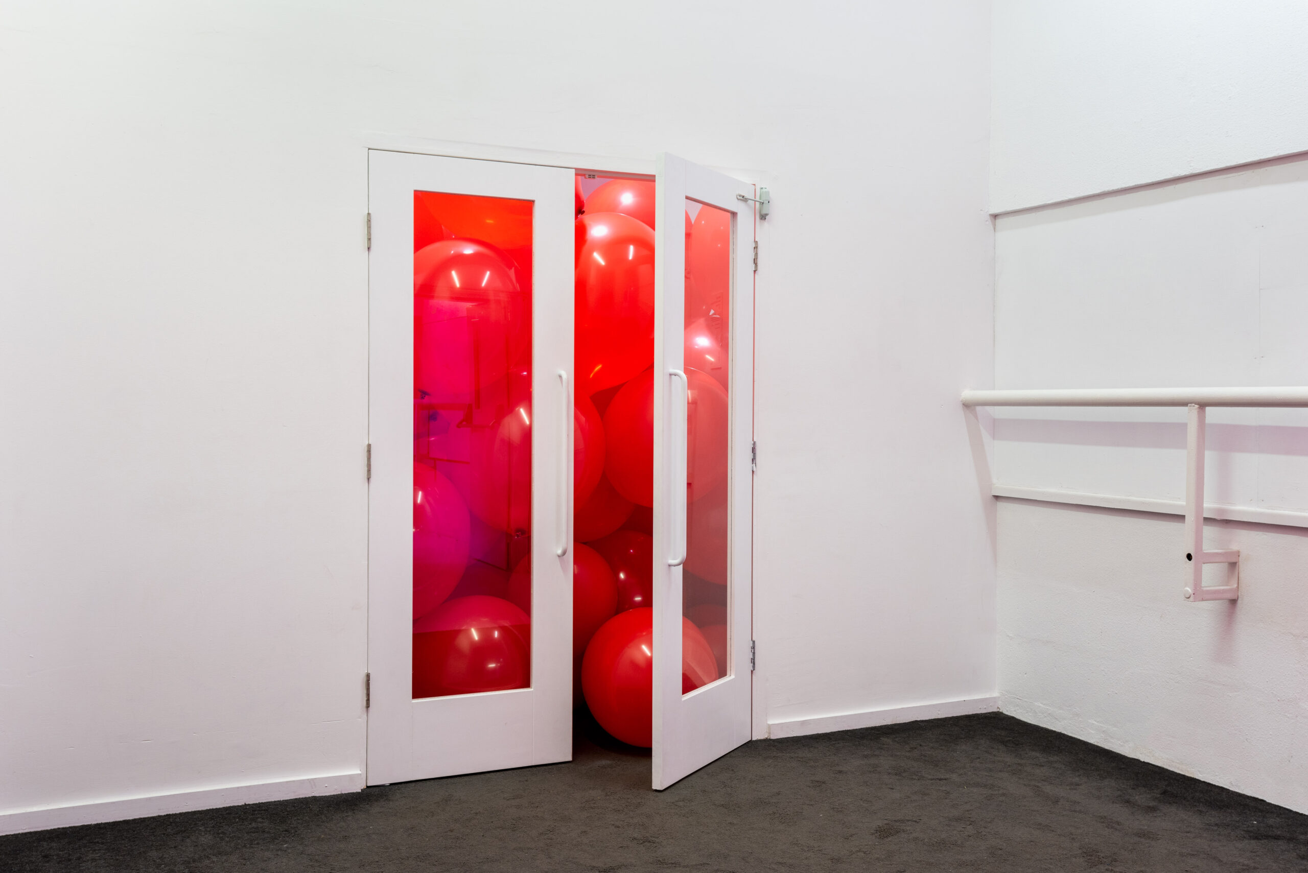 ID: a pair of glass double-doors are left slightly ajar on an expansive white wall. Behind the glass doors there are hundreds of large red balloons that completely fill the view into the room.