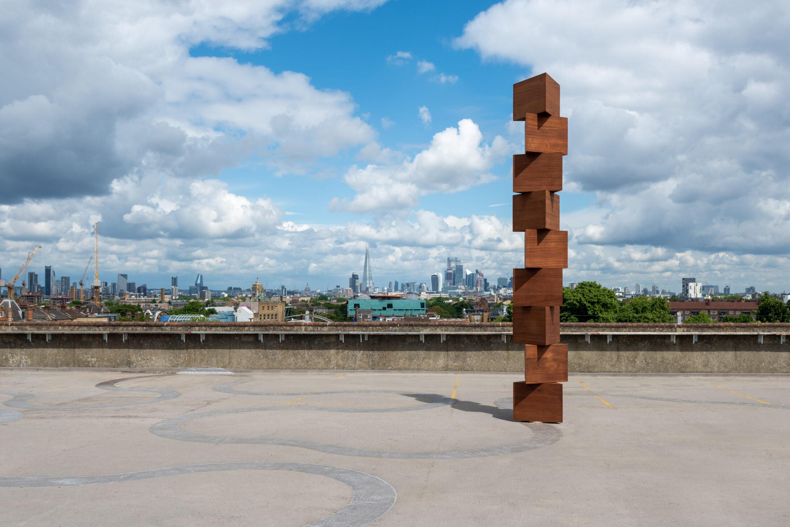 ID: a large wooden obelisk is made from nine rotating triangular prisms stacked on top of one another and rotating to create a bird’s eye view of a nine-pointed star. The sculpture measures over 4.5 metres in height and is made from African Mahogany. It is placed on a concrete rooftop with the London skyline and clear blue skies in the background.